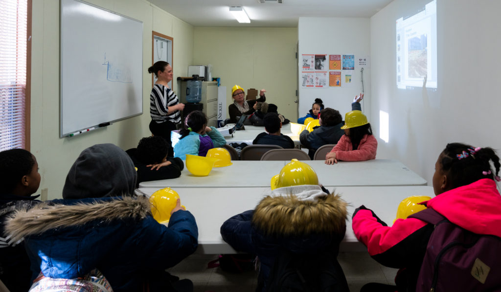 Students wearing yellow hard hats sit in a room and listen to a woman talk about the construction of Essex Crossing